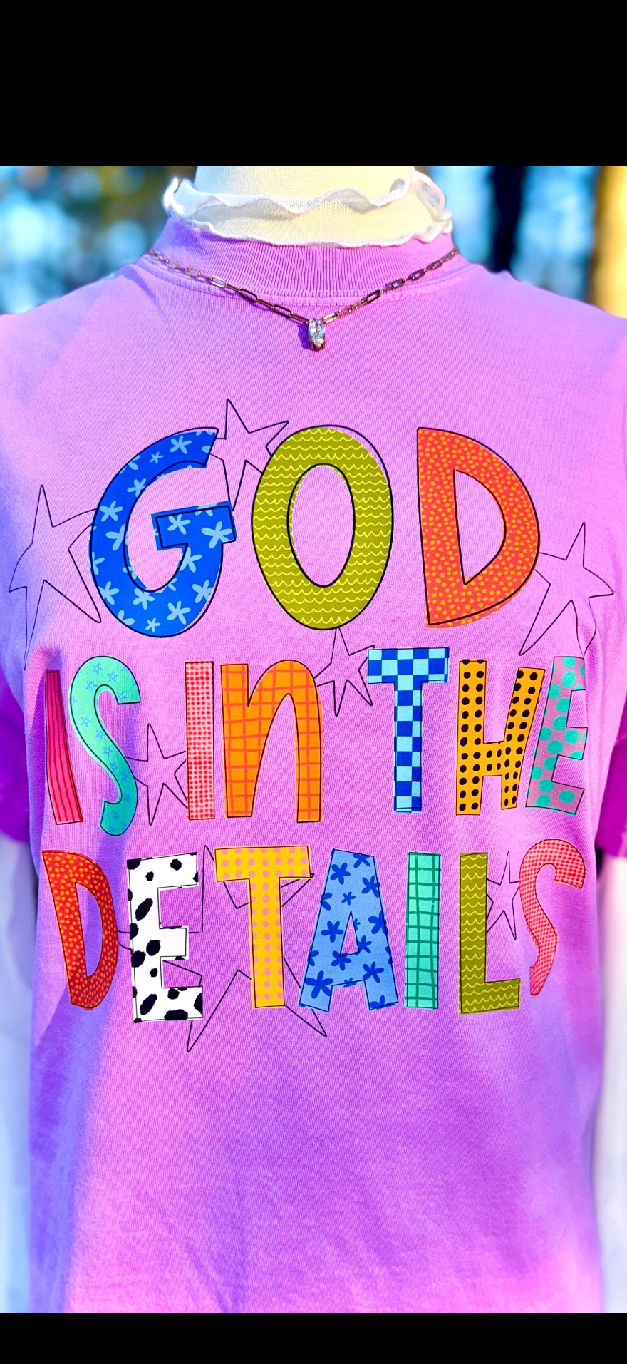 Purple shirt with colorful text 'GOD IS IN THE DETAILS' on a blurred background