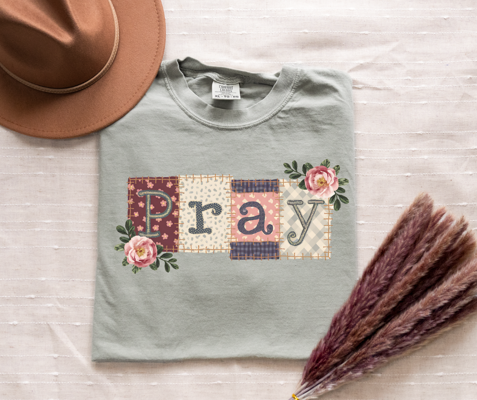 Gray sweatshirt with 'Pray' design on a wooden surface with a hat and dried flowers.