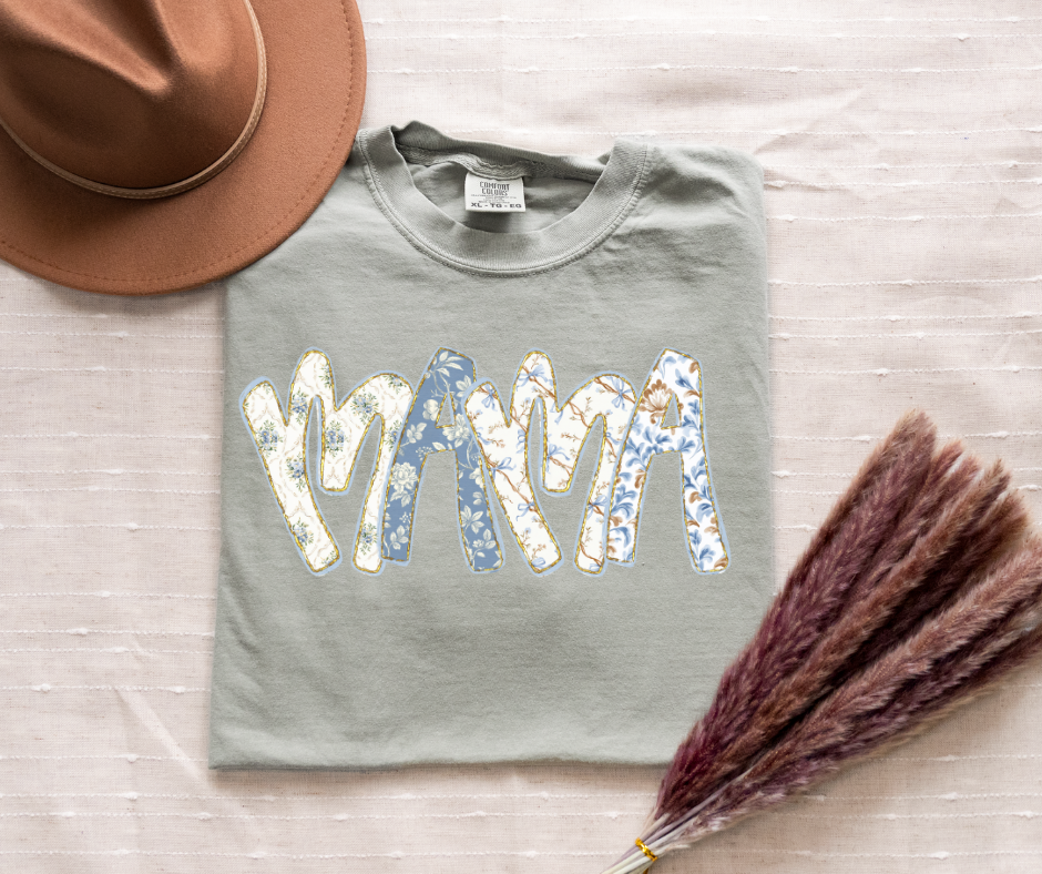 Gray sweatshirt with 'MAMA' text on a light wooden surface with a hat and dried plant.