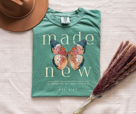 Green t-shirt with butterfly design and text, placed on a light surface with a hat and dried flowers.
