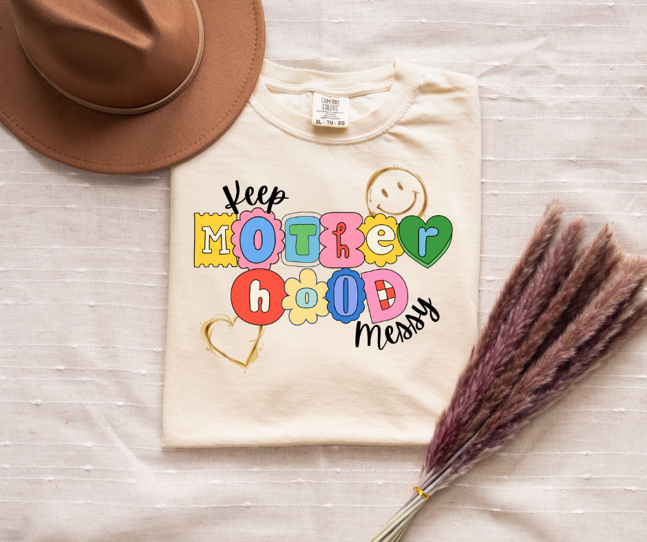 Beige t-shirt with colorful text design on a wooden surface with a hat and dried plants.